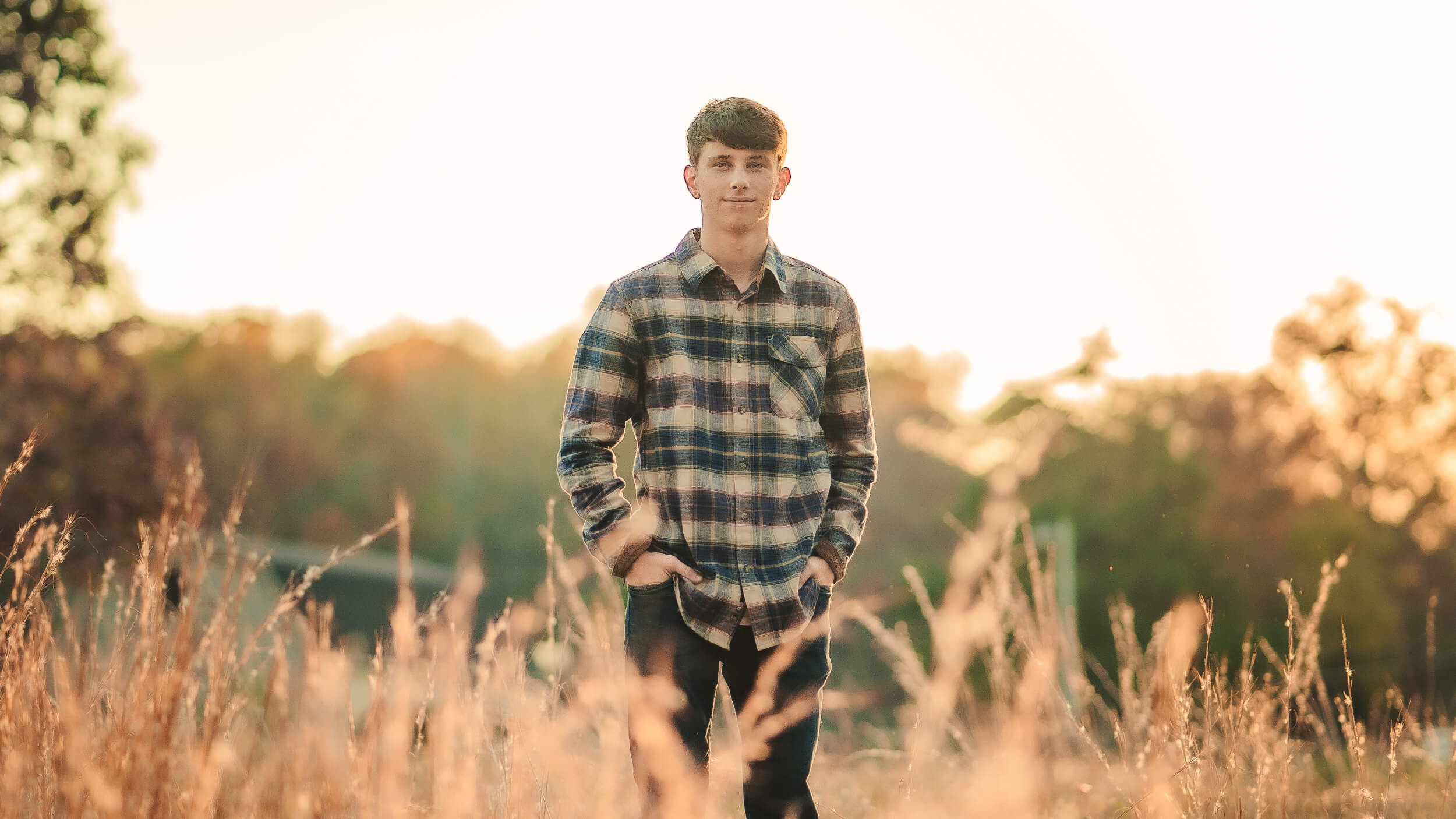 A young man - Dylan Michaud - in a plaid shirt and blue jeans standing in a field with with tall brown grass at sunset.